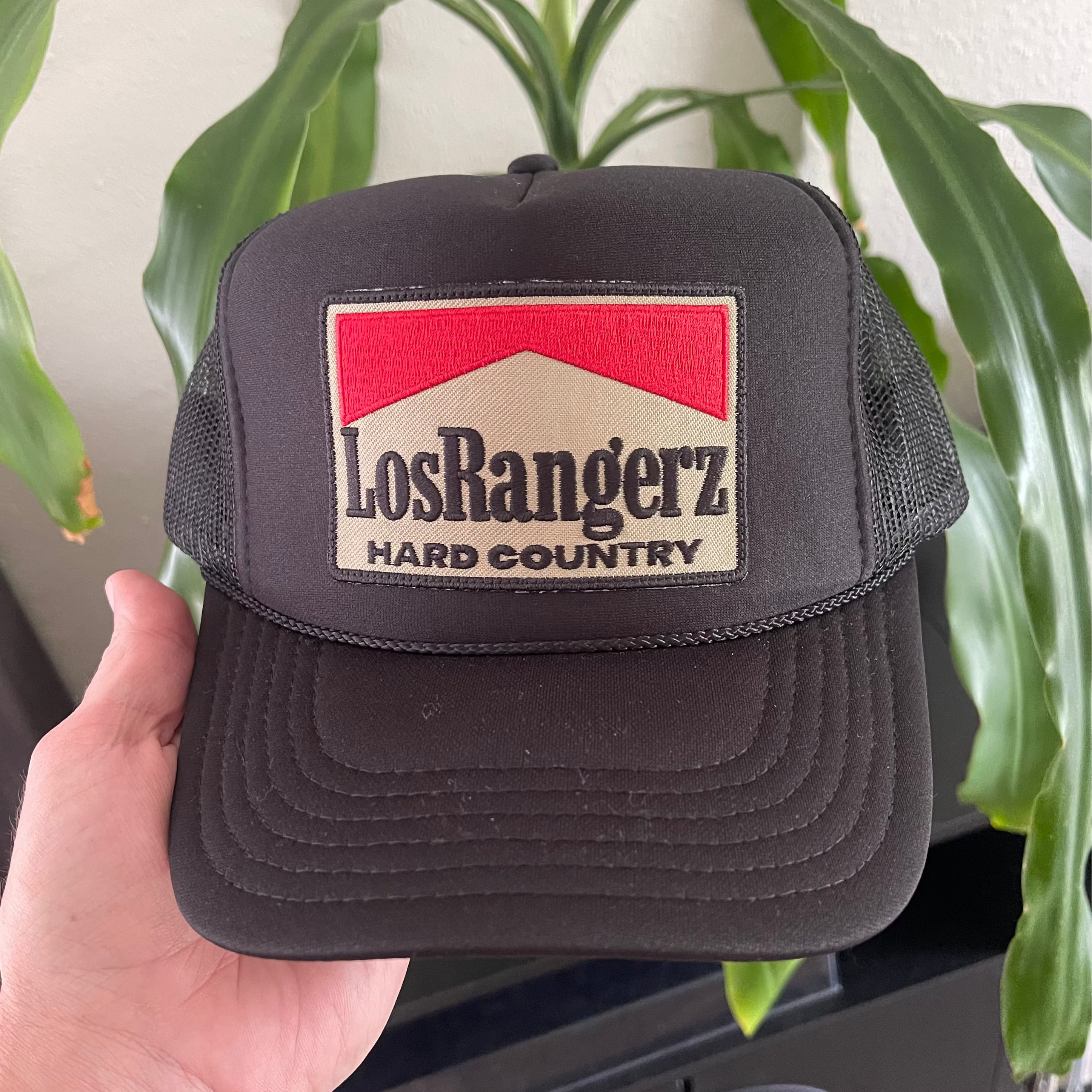 Black trucker hat with 'Los Rangerz Hard Country' logo held in front of a plant.