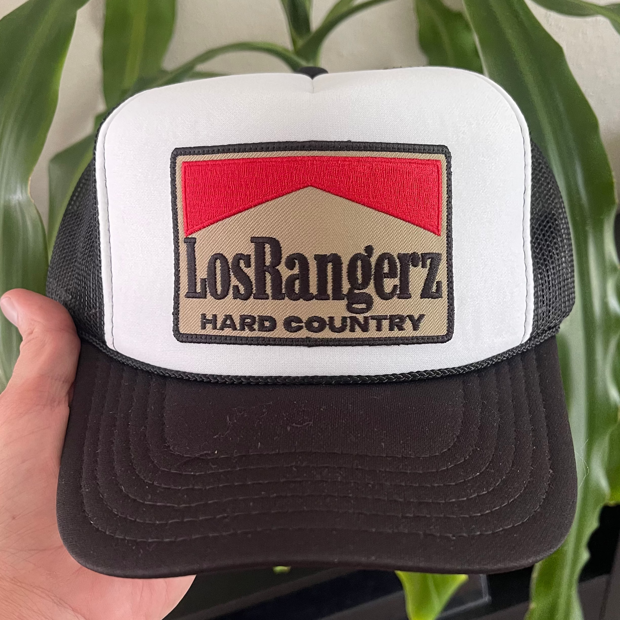 Black and white trucker hat with 'Los Rangerz Hard Country' logo held by a hand against a plant background.