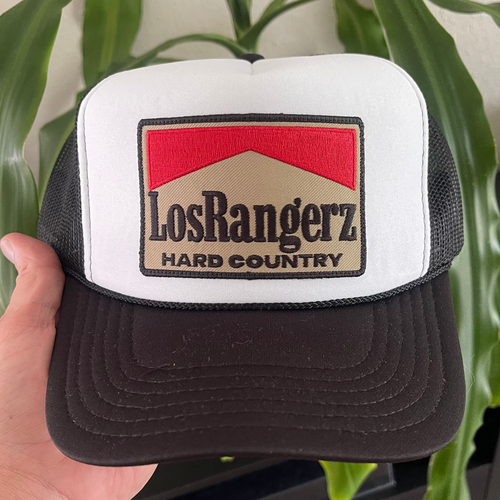 Black and white trucker hat with 'Los Rangerz Hard Country' logo held by a hand against a plant background.